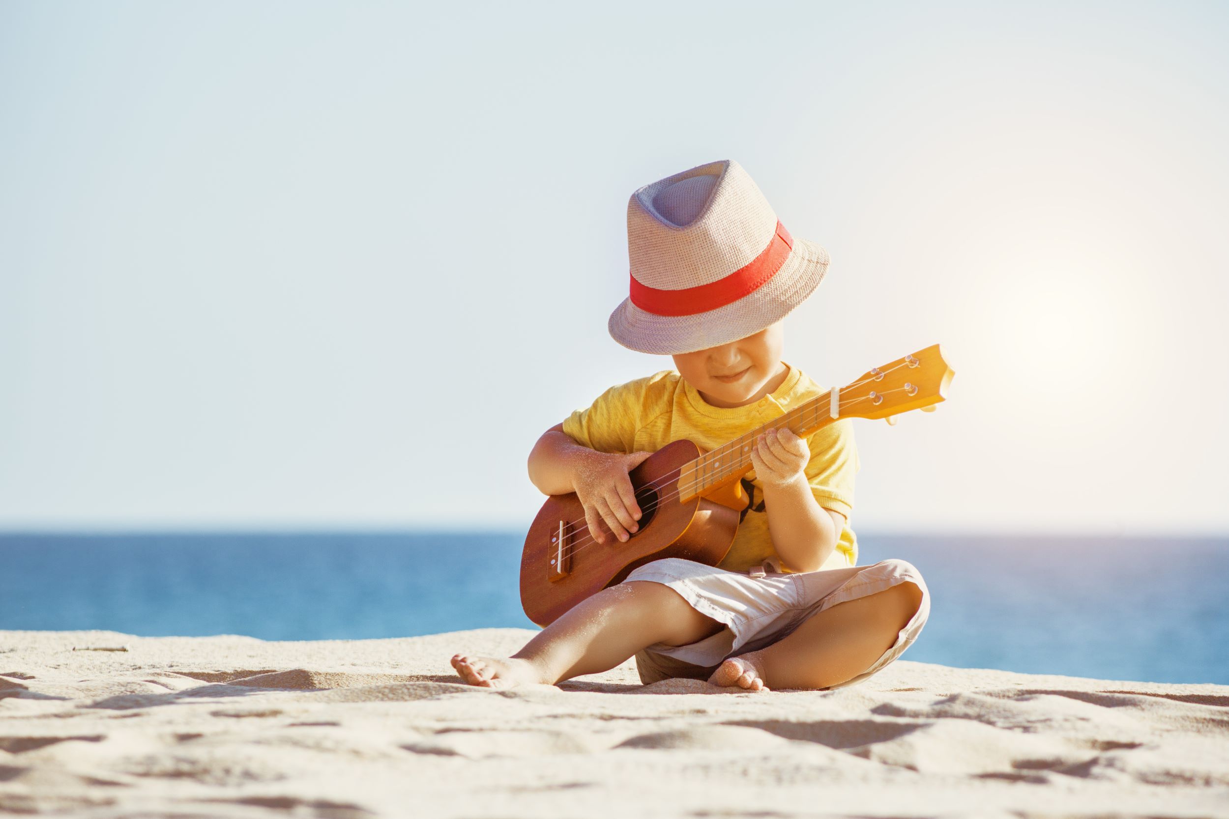 Kleiner Junge mit Sonnenhut spielt Ukulele am Strand – Probestunde vor dem Schuljahresstart in der Musikschule Einfach Musikalisch Erfurt