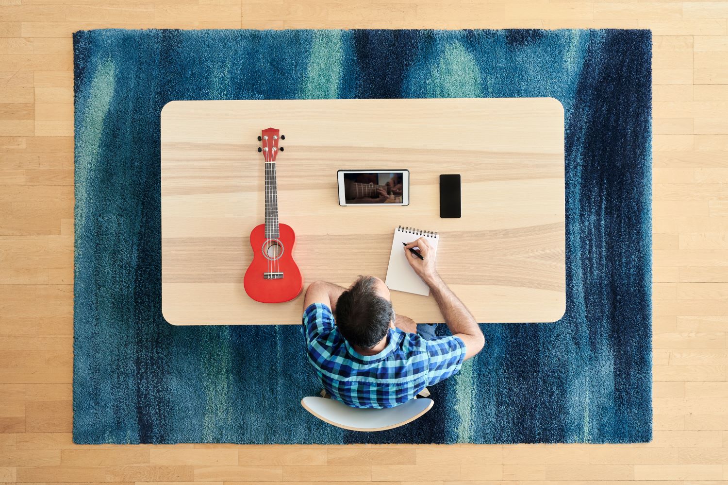 Ein Mann sitzt am Tisch, neben ihm liegen eine rote Ukulele, ein Tablet und ein Handy – von oben fotografiert